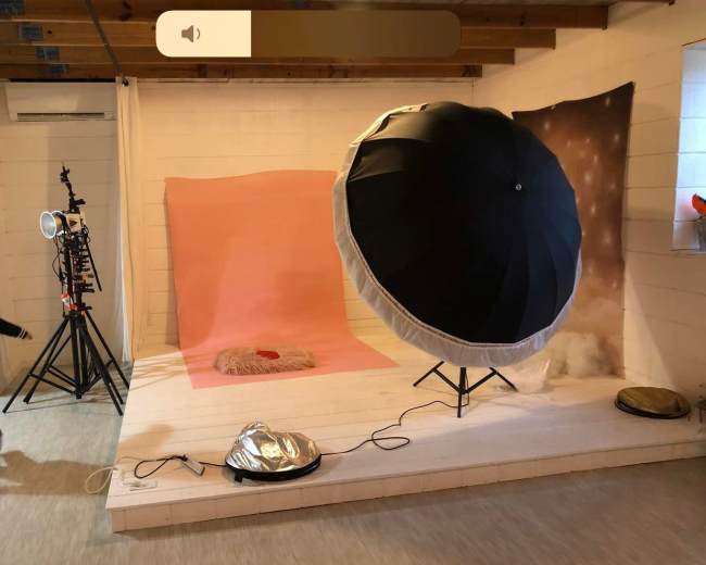 A photography setup with a large umbrella softbox, backdrops in shades of pink and neutral tones, and various tripod equipment arranged in a studio space.
