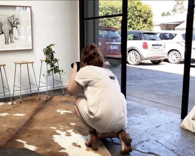 A person is crouching on a cowhide rug while taking a photo of a framed illustration of an elephant in a well-lit room.