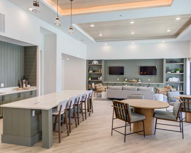 The image shows a modern, open-concept lounge area with a kitchen counter, seating arrangements, and wall-mounted televisions.