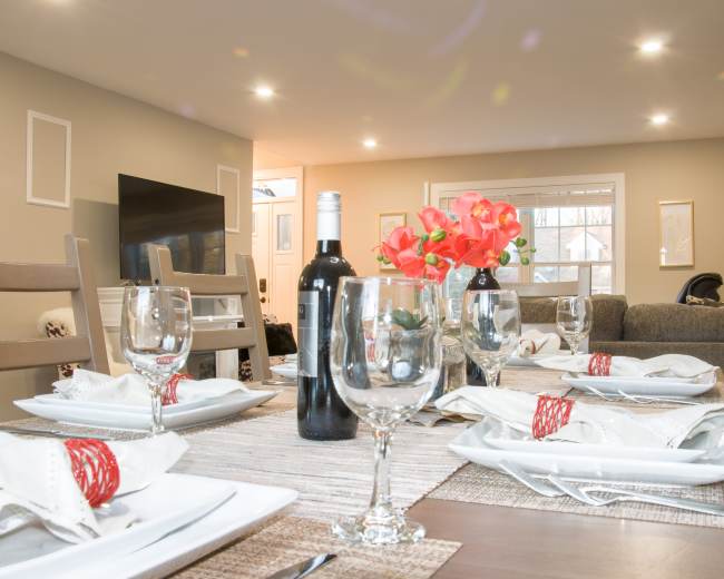 A dining table is set with wine bottles, glasses, and neatly arranged plates, while a cozy living area with a television is visible in the background.