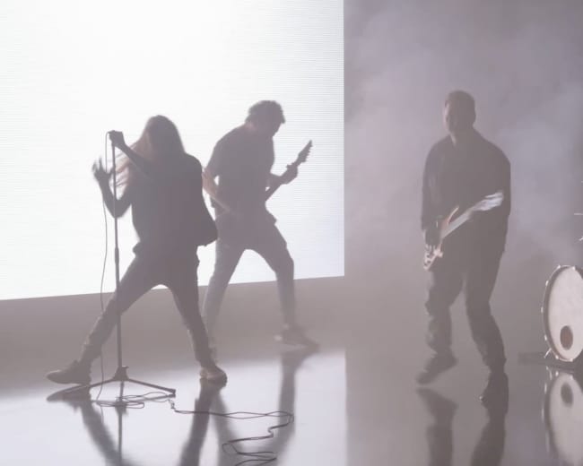 A band performs on stage in front of a bright screen, with fog surrounding them and reflections visible on the floor.