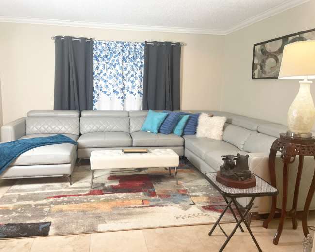 The living room features a gray sectional sofa, a decorative rug with colorful patterns, and a small side table beside a lamp.