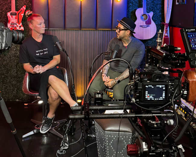 A man and a woman are sitting in a recording studio with musical instruments and cameras set up for an interview.