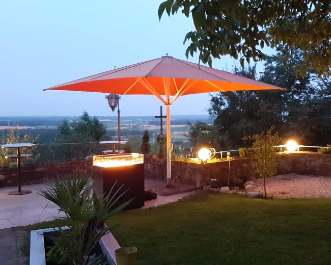 A patio scene features an orange umbrella over a grill, surrounded by landscaped greenery and glowing orbs of light in the dusk.