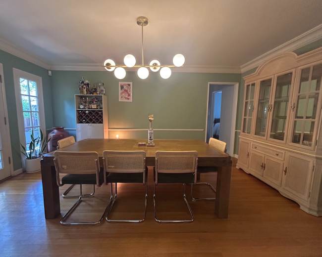 The image shows a dining room with a large wooden table surrounded by four chairs, an open shelving unit, and a cabinet against a green wall, illuminated by a modern chandelier.
