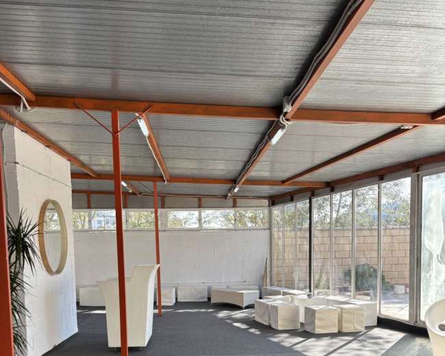 The image shows a spacious, modern room with a gray carpet, large windows, and minimal furniture, including white seating and a circular mirror on the wall.