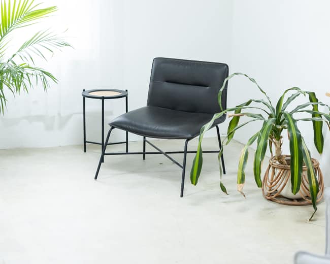 A black chair and a small side table are positioned in a bright corner of a room with white walls and a large window.