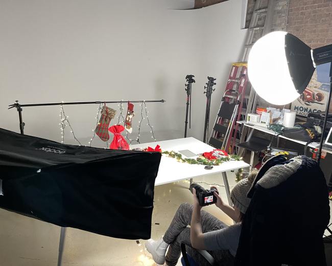 A person sits in a chair holding a camera in a studio setup with a white backdrop, holiday decorations, and lighting equipment arranged for a photo shoot.