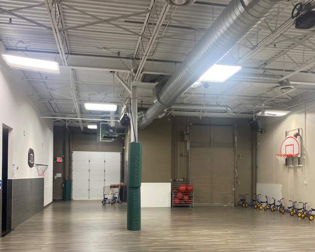 The image shows a spacious indoor gym with basketball hoops, exercise equipment, and a hardwood floor.