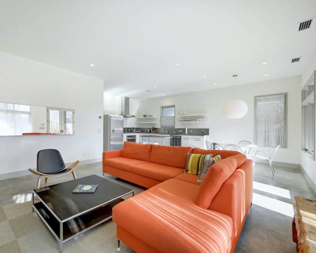 The image shows a modern living area with an orange sectional sofa, a black coffee table, and a view of a kitchen in the background.