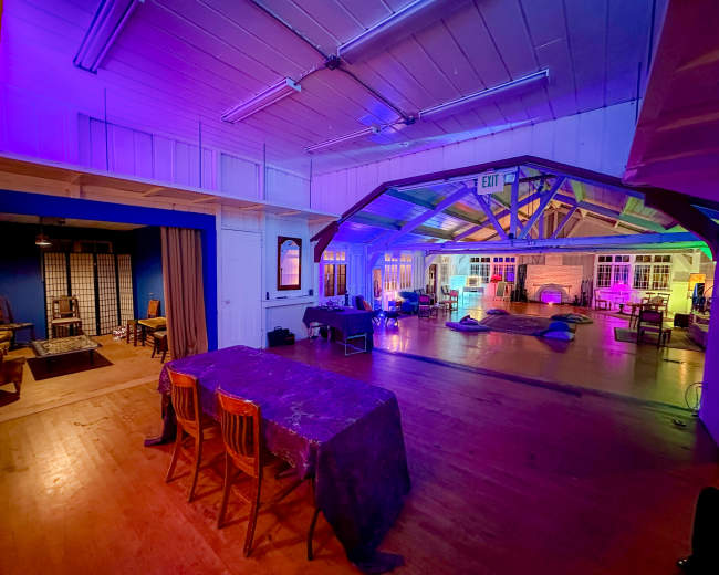 The image depicts a spacious indoor area featuring a wooden floor, multiple seating arrangements, and colorful lighting that illuminates the walls and ceiling.