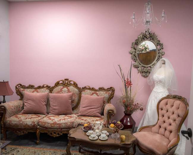 A room features a pink wall, vintage furniture with floral upholstery, and a wedding dress displayed on a hanger beside a mirror.