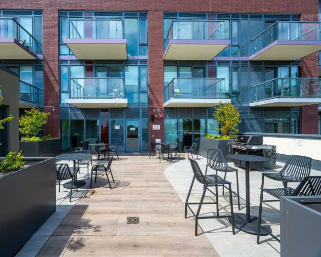 The image shows a modern outdoor patio area with tables and chairs in front of an apartment building featuring multiple balconies.
