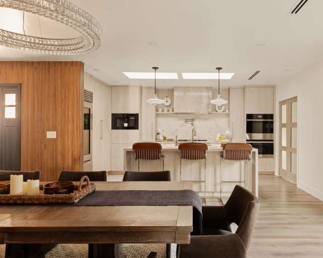 The image shows a modern kitchen and dining area with a wooden dining table, stylish seating, and a kitchen island featuring pendant lighting and a marble backsplash.