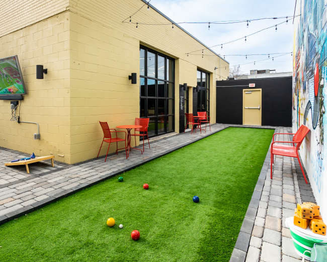 A courtyard features a green lawn with bocce balls, red chairs and tables, and a TV mounted on a yellow wall.