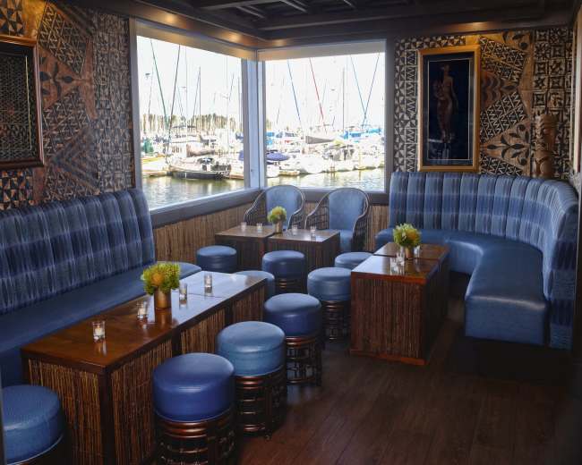The image depicts a cozy waterfront lounge area featuring blue upholstered seating, wooden tables, and a view of sailboats through large windows.