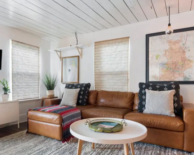 The image shows a cozy living room featuring a brown leather sofa, a round table, a wall-mounted television, and plants near large windows with bamboo shades.