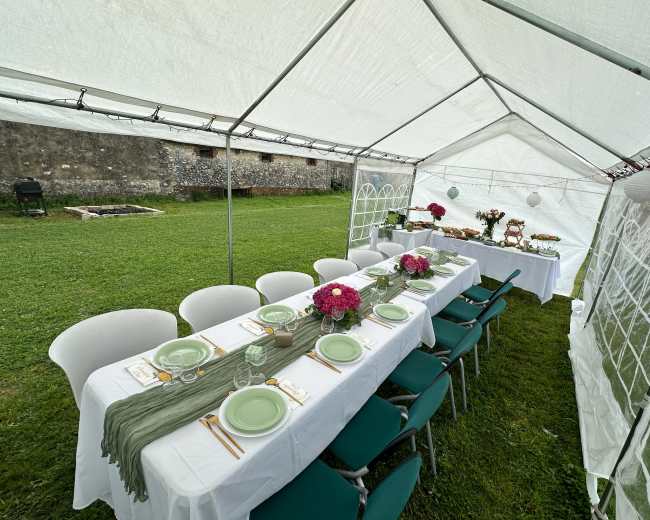 A long table set with plates and floral arrangements is prepared under a white tent on a grassy area.