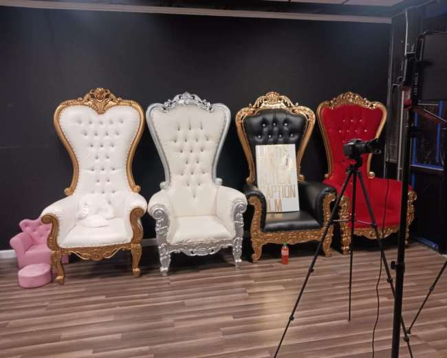 The image displays four ornate, high-backed chairs with different colors and designs set against a dark backdrop, accompanied by a camera on a tripod.