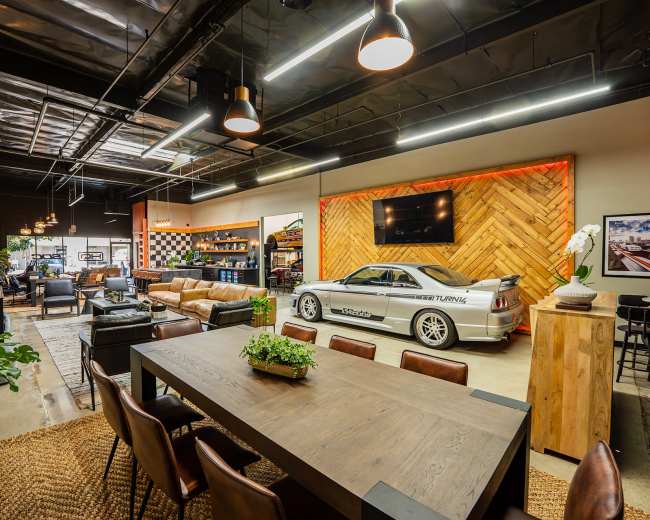 The image shows a modern, industrial-style lounge area featuring a sports car, large seating arrangements, and a dining table, with mixed decor elements including wood and plants.