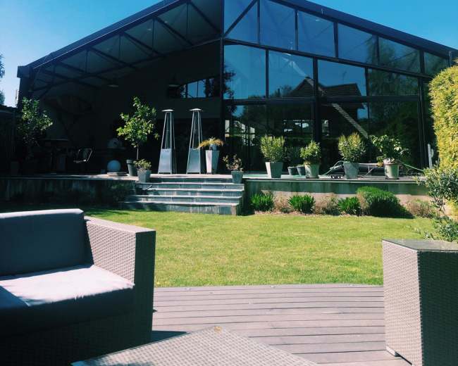 A modern glass-panel house with a sloped roof is surrounded by well-maintained greenery and outdoor seating.