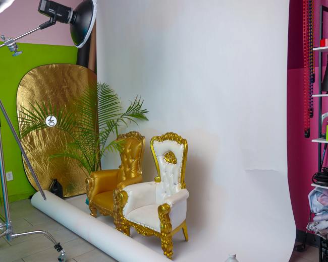 The image shows a photography studio setup with two ornate chairs, one gold and one white, in front of a white backdrop.