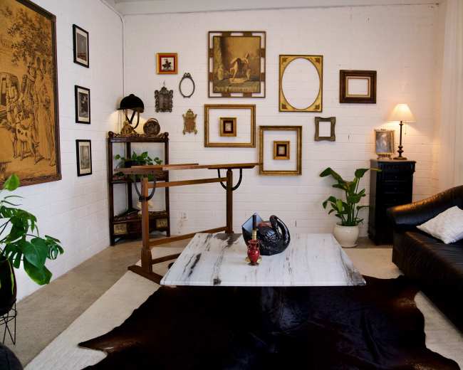 The image shows a stylish living room with a black leather sofa, a coffee table on a cowhide rug, various framed artworks on the wall, and a lamp beside a tall plant.