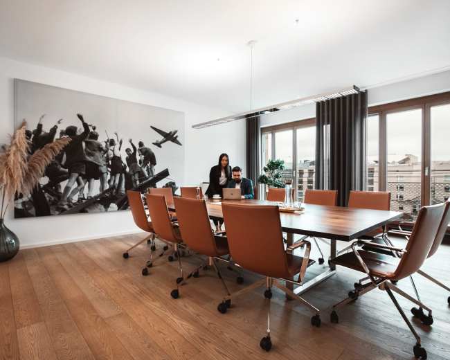 A modern conference room features a large wooden table surrounded by orange chairs, with a black-and-white mural on the wall and a group of people engaged in discussion.