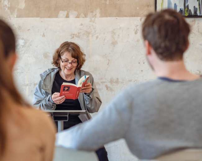 A woman reads from a book while seated at a podium in front of an audience.