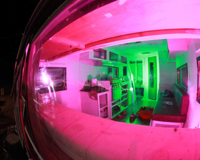 A brightly lit interior of a small structure features green and pink lighting, showcasing shelves and a seating area.