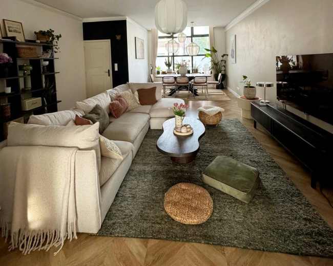 The image shows a spacious living room with a light-colored sectional sofa, wooden coffee table, and a television unit, all arranged around a large area rug.
