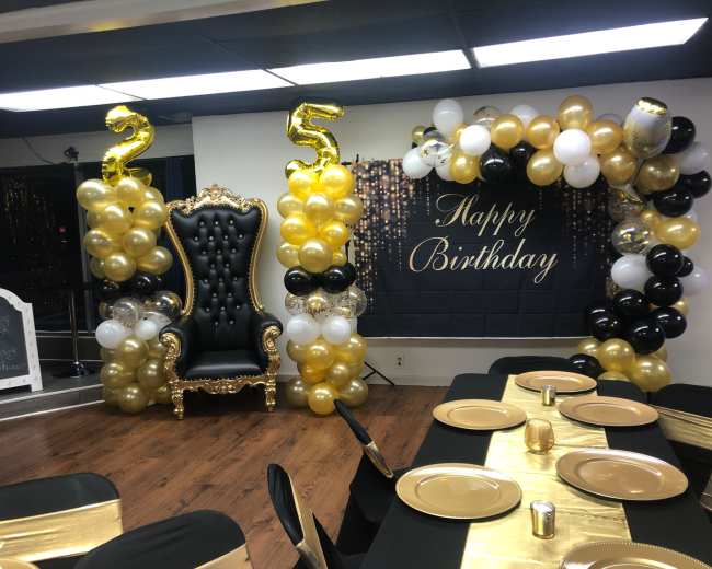 A birthday celebration setup featuring a black and gold color scheme, with a large throne chair, an arch of balloons, and a "Happy Birthday" banner.