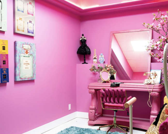 The image shows a vibrant pink room featuring a vanity desk with a mirror, a plush blue rug, and decorative elements on the walls.