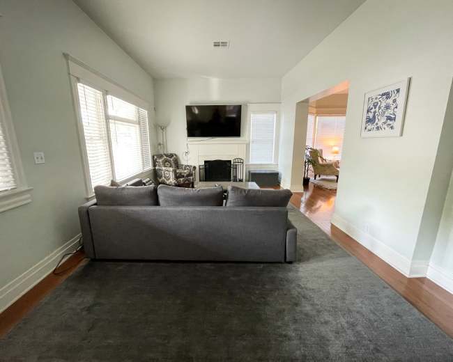 A living room with a gray sofa facing a wall-mounted television, situated near windows with white blinds.