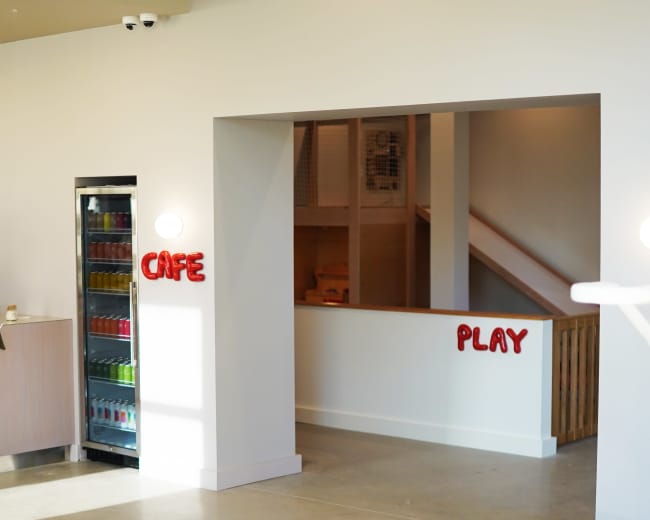 The image shows a bright interior space with a "Café" area on the left and a "Play" area beyond an open entrance on the right.