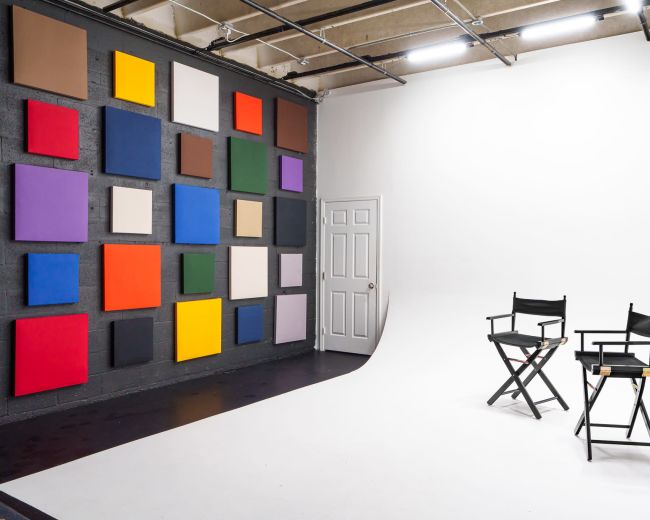 A photo studio features a wall with colorful canvases and a white backdrop with two director's chairs.