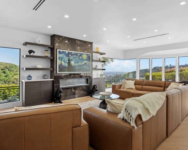 The image features a modern living room with large windows, a sleek fireplace, and brown leather couches arranged around a glass coffee table.