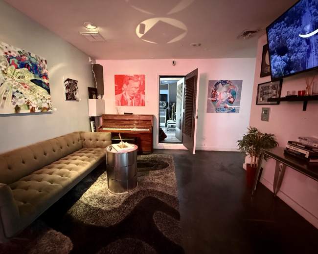 The image shows a modern lounge area with a tufted sofa, a round table, a piano, colorful artwork on the walls, and a doorway leading to another room.