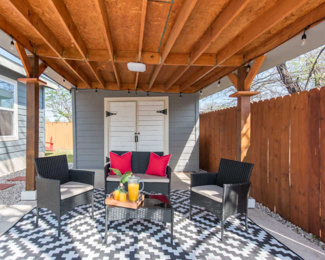 The image shows a covered outdoor seating area with four chairs arranged around a small table, set on a patterned rug beneath wooden beams.