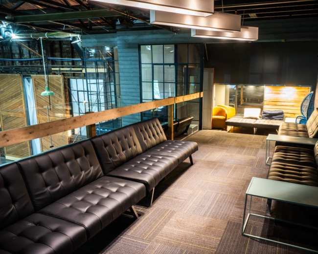 The image shows a modern lounge area with dark leather couches, wooden walls, and various seating options, illuminated by overhead lighting.