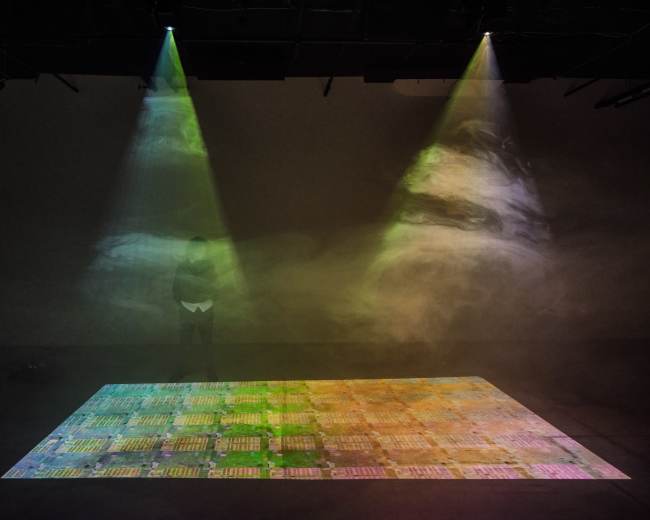 A person stands on a projection that displays a colorful, patterned surface illuminated by beams of light from above.