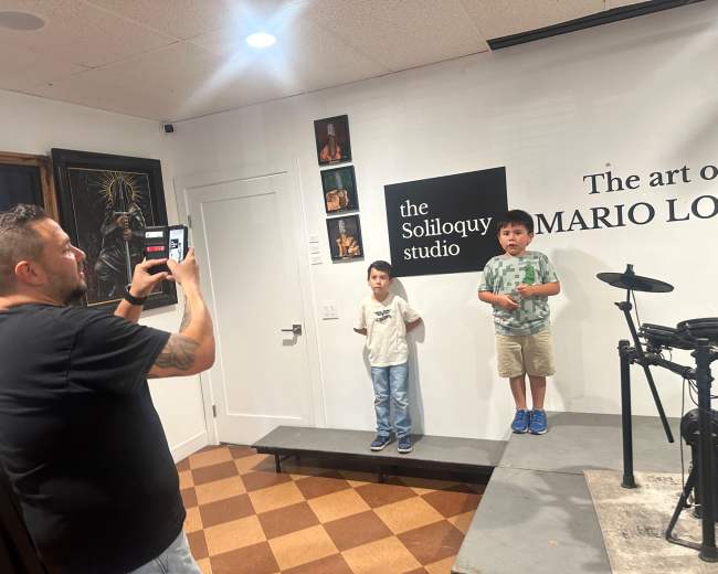 A man is taking a photo of two boys standing on a small stage in an art studio, with gallery walls displaying various artworks.