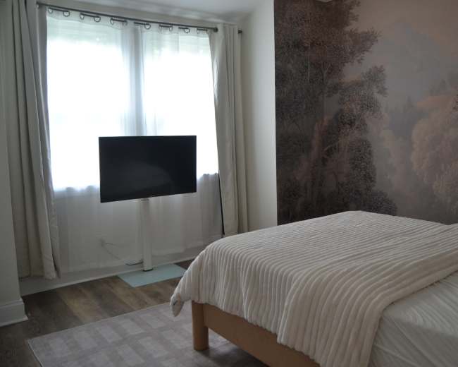 The image shows a simple bedroom with a bed covered in a light blanket, a television on a stand, and a large window covered by sheer curtains.