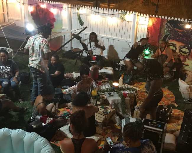 A group of people gathers on a lawn, sitting on blankets and chairs, while enjoying a nighttime party illuminated by string lights.
