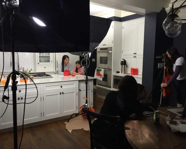 A woman and a child are engaged in an activity at a kitchen island while a crew sets up filming equipment in the background.