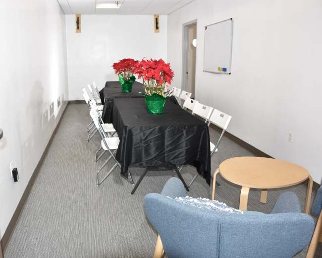 A meeting room is set up with a black tablecloth over a long table flanked by white chairs, featuring two green vases of red poinsettias.