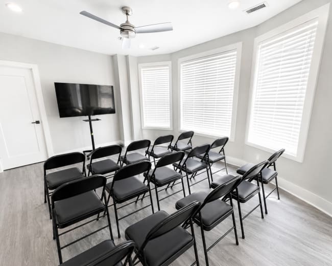 The image shows a lightly furnished meeting room with several rows of black folding chairs facing a wall-mounted television and large windows with blinds.