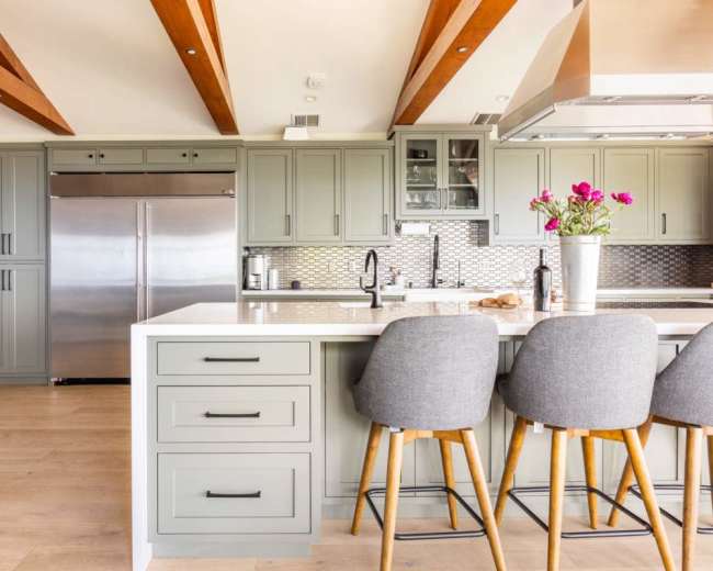 The image shows a modern kitchen with light green cabinets, stainless steel appliances, and a central island with wooden bar stools.