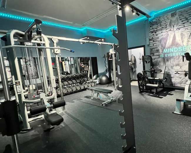 The image shows a well-equipped gym featuring various fitness machines, free weights, and a motivational wall mural.
