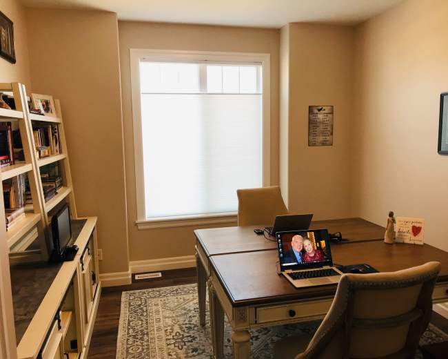 A small, light-colored office features a wooden desk with two laptops open, a bookshelf filled with books, and a window with shades letting in natural light.